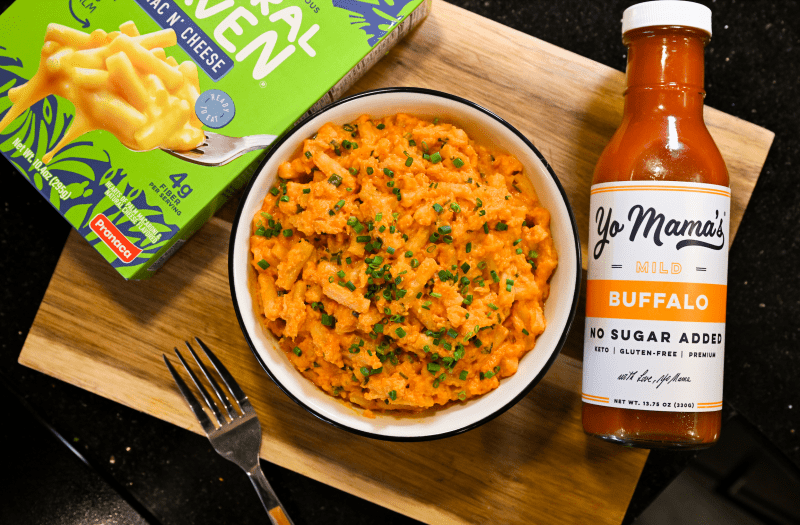 Bowl of creamy buffalo-style hearts of palm pasta topped with chopped chives, served on a wooden board beside a box of Natural Heaven mac and cheese and a bottle of Yo Mama’s mild buffalo sauce.